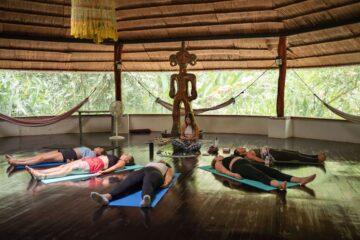 In a spacious yoga studio with natural light flooding in through large windows, a group of individuals are laying down in what appears to be the Savasana pose (corpse pose) on yoga mats.